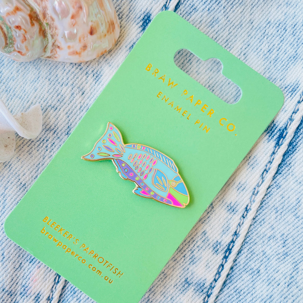 Colorful Parrotfish shaped enamel pin on a green card with 'Braw Paper Co' branding, placed on a textured fabric background.