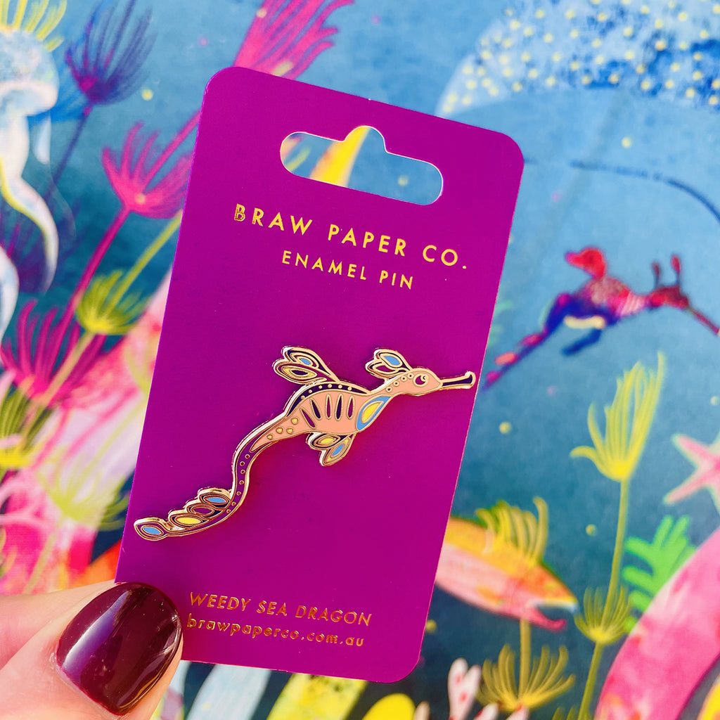 Weedy sea dragon enamel pin on a purple card with 'Braw Paper Co.' branding, against a colorful underwater illustration background.