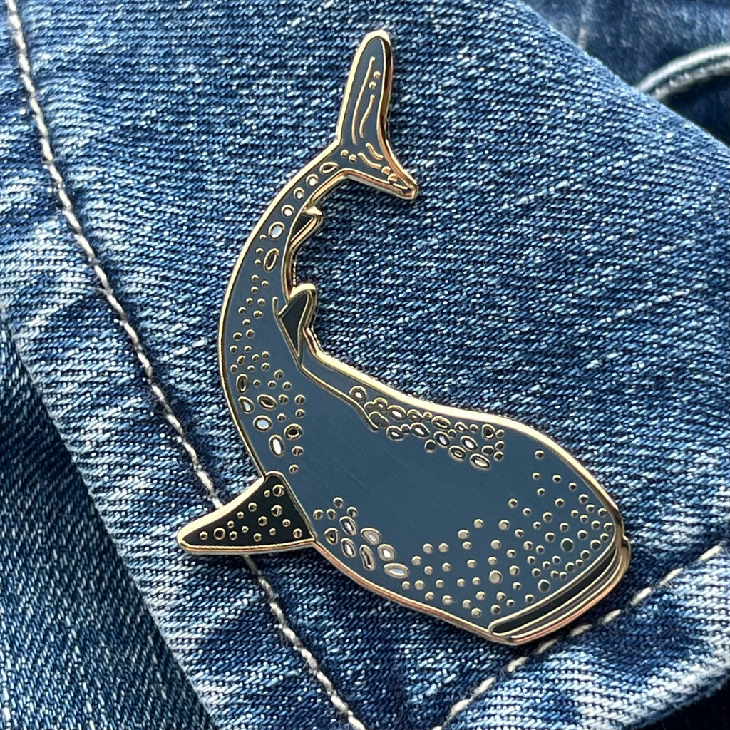 An enamel pin shaped like a whale shark, with intricate details, placed on denim fabric.