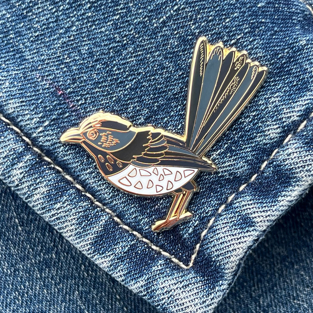 An enamel pin shaped like a Willie Wagtail bird, with black and white plumage, attached to a denim fabric.