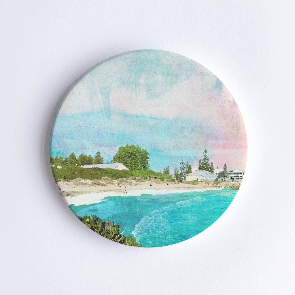 Round, hand printed ceramic coaster with illustration of Bather's Beach in Western Australia with turquoise water, sand dunes, buildings and trees.