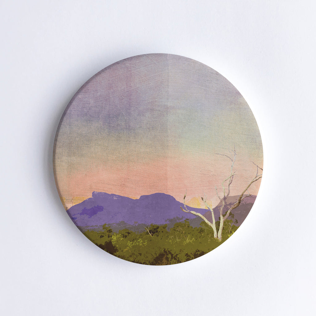 Round hand printed ceramic coaster with illustration of a purple shining Bluff Knoll mountain during sunset with a bare tree and trees in the foreground.