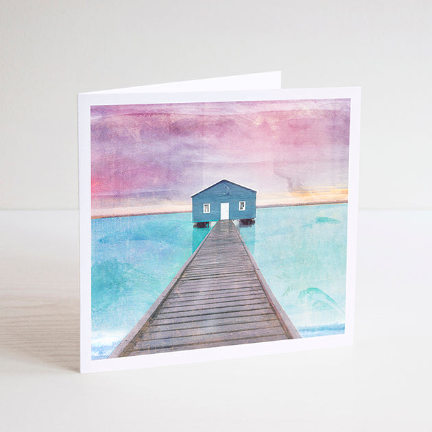 Square notecard with illustration of the blue Crawley Boathouse at the end of the pier with turquoise water and pink sky. 
