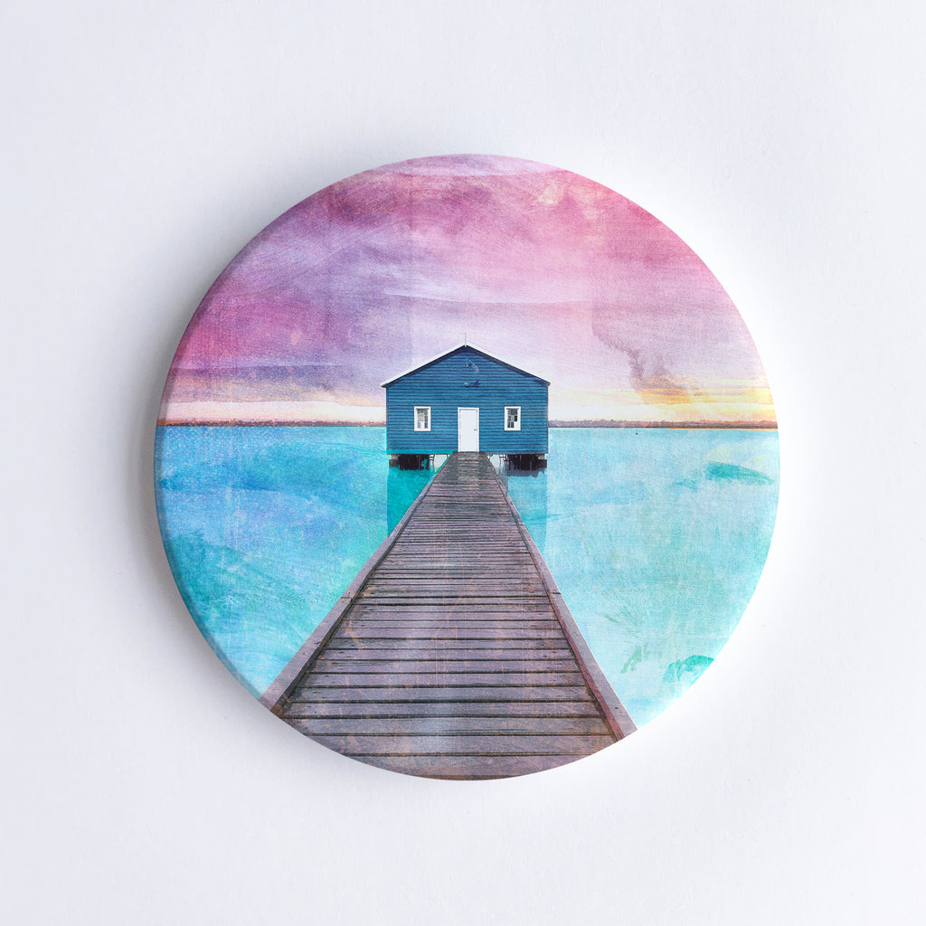 Round, hand printed ceramic coaster with illustration of the blue Crawley Boathouse at the end of the pier with turquoise water and pink sky. 