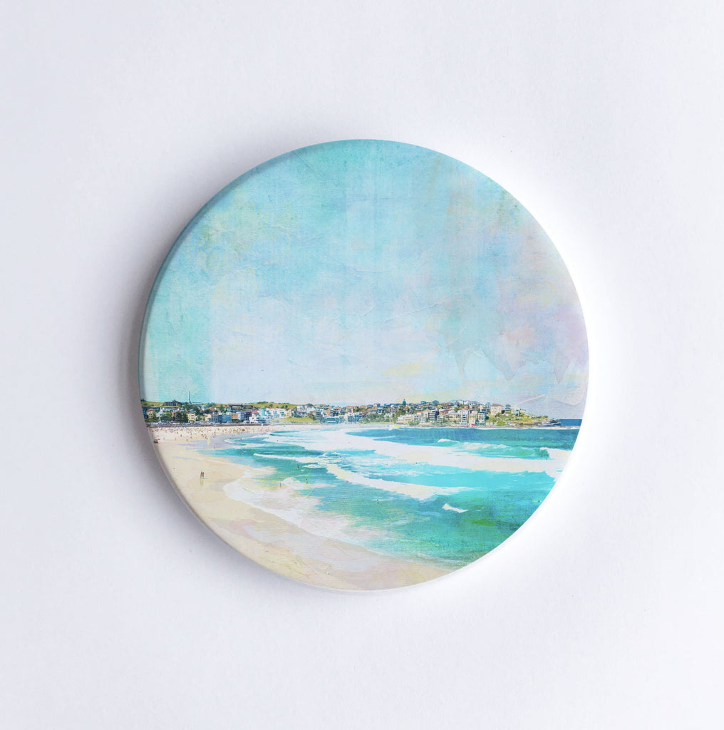 Round, hand printed ceramic coaster with illustration of Bondi Beach in Sydney showing waves and houses on a hill in the background.