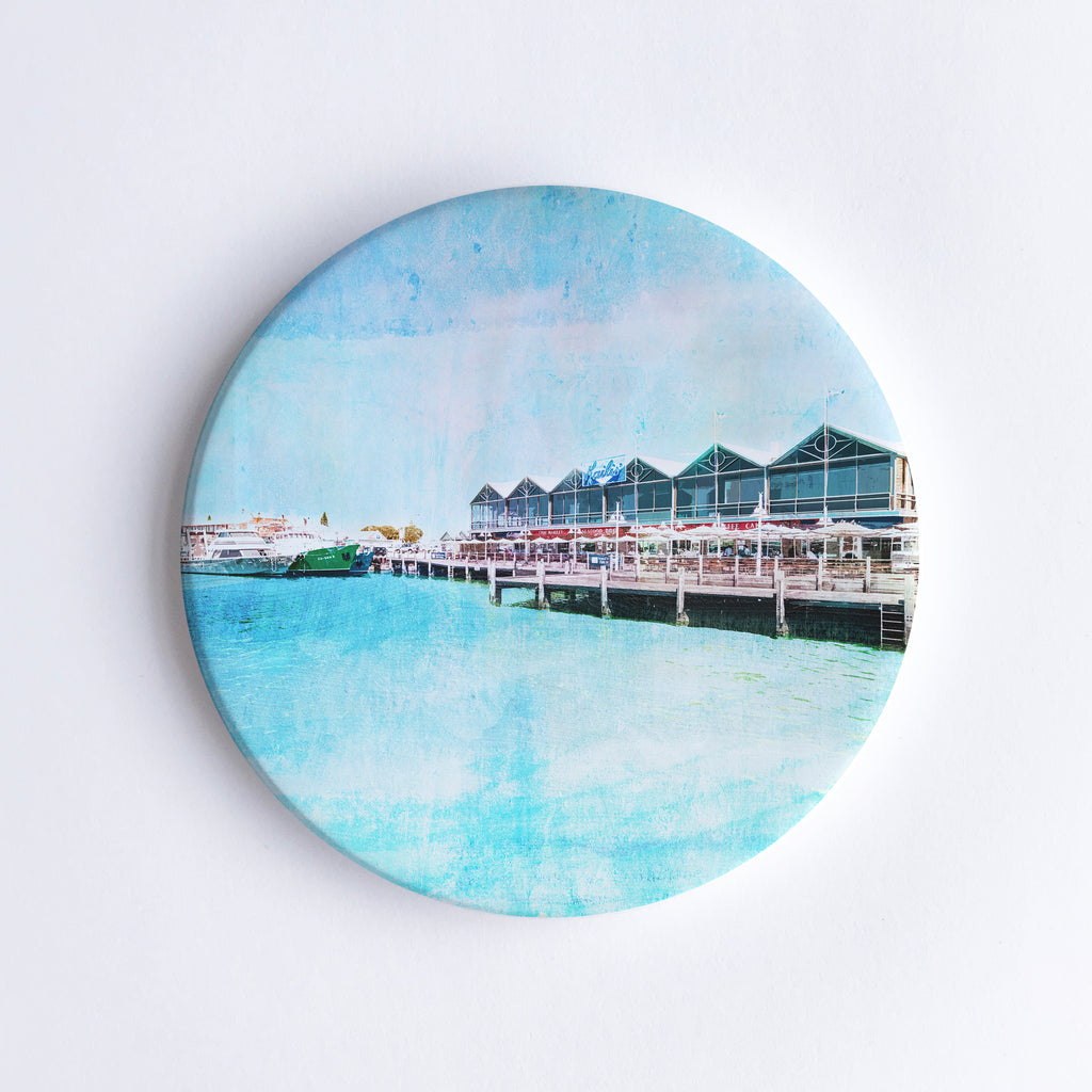 Round, hand printed ceramic coaster with illustration of Fremantle Fishing Boat Harbour and Kaili’s fish restaurant on stilts over the water.