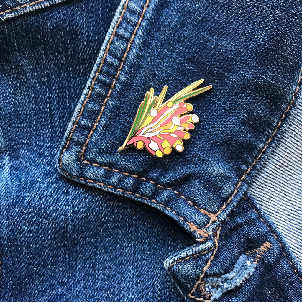 Enamel pin with red, yellow, orange, green and white kangaroo paw flowers illustration.