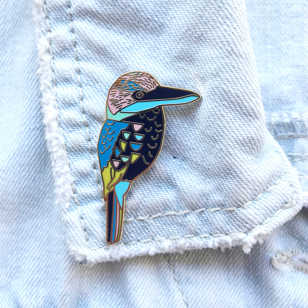 An enamel pin depicting a Blue-Winged Kookaburra bird, attached to a denim fabric.