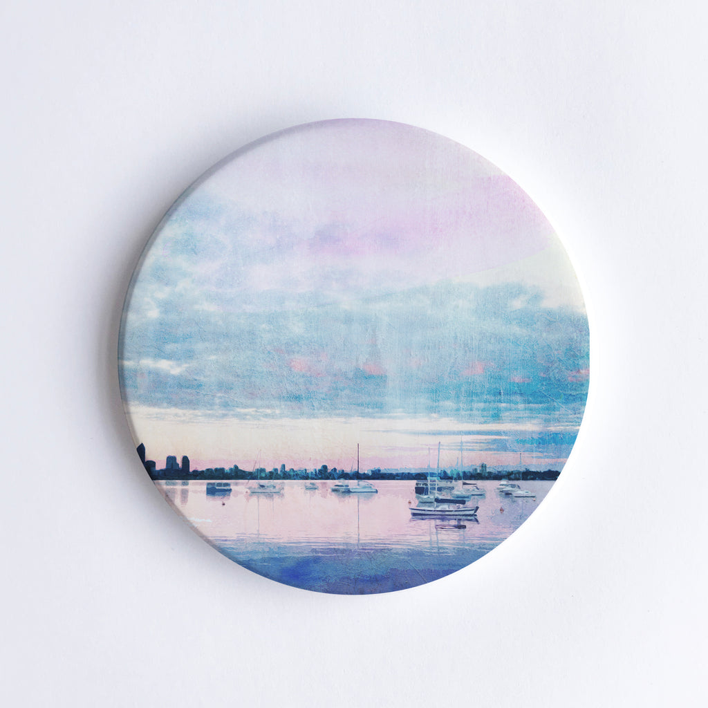 Round, hand printed ceramic coaster with illustration of Matilda Bay at dusk, sailing boats on Swan River and Perth city skyline in the background