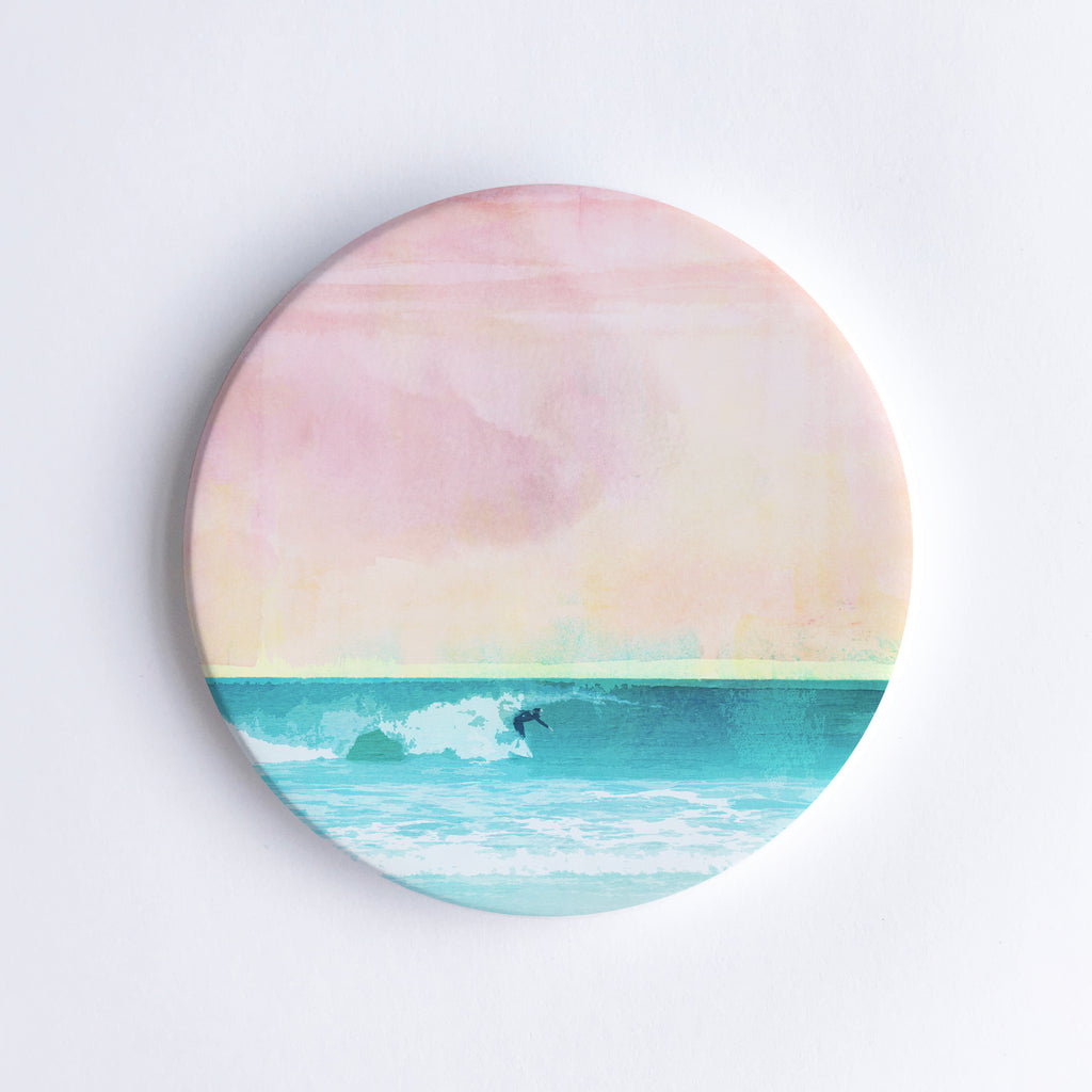Round ceramic coaster with  an illustration of a solo surfer riding a wave at sunset.